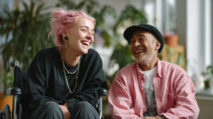 A young woman with pink hair and an older man in a wheelchair share a joyful moment of laughter.